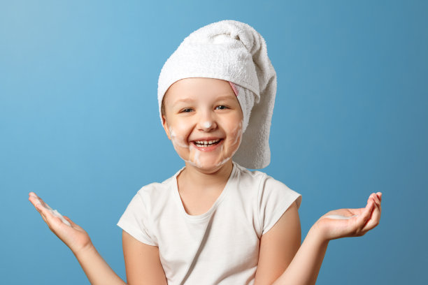 小女孩头上裹着一条白毛巾，在蓝色的背景下洗头。孩子笑了，在脸上涂了泡沫。日常卫生的概念。图片下载