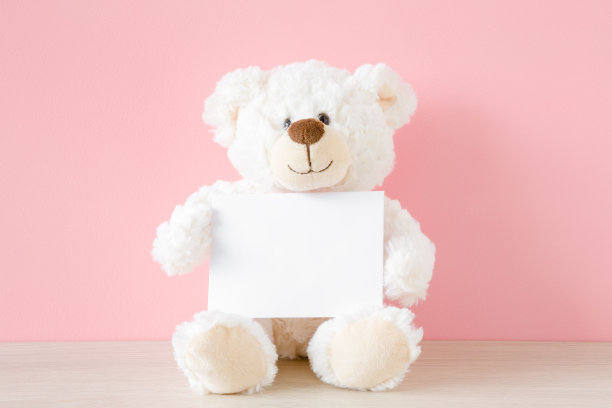 微笑的泰迪熊坐在淡粉色的墙上，拿着白色的卡片。空的地方，为灵感，情绪，感伤的文本，可爱的引用或语录好心情。前视图。图片下载