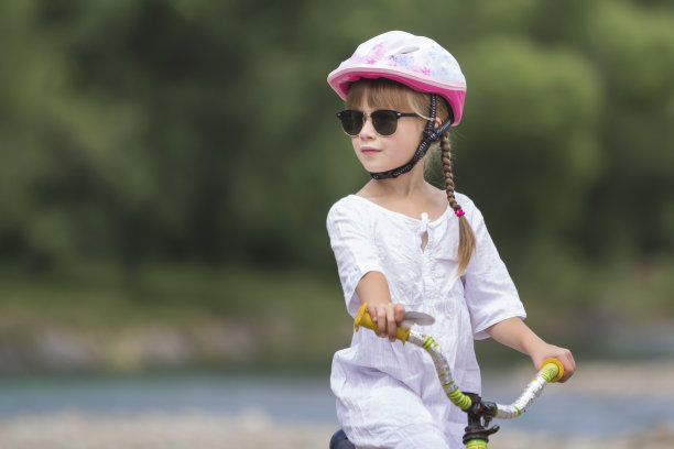 特写照片的骄傲漂亮的年轻女孩在白色的衣服，太阳镜与长金黄色的辫子戴粉红色的安全帽骑在模糊的绿树夏天拷贝空间背景儿童自行车。图片下载