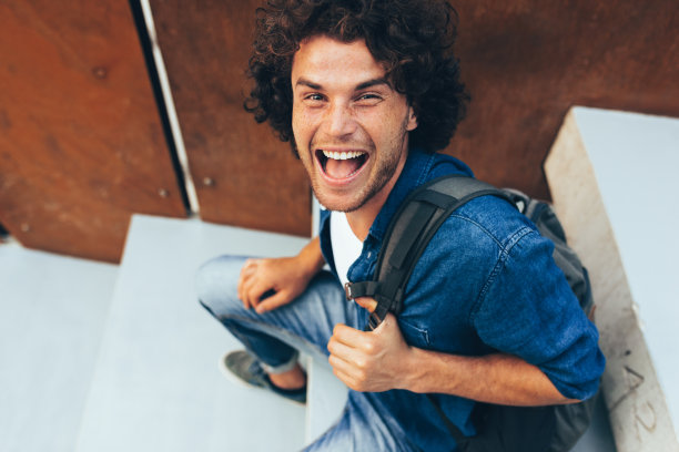 一个快乐的年轻人面带微笑，坐在城市街道的混凝土楼梯上，背上背着背包。积极的学生男生穿着休闲装，在校园户外放松。人的生活方式图片下载