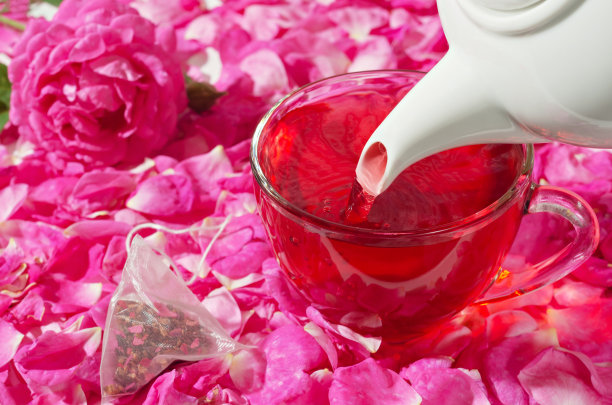 一杯热腾腾的花茶从茶壶里倒出来，旁边是一个茶包和玫瑰花瓣。有选择性的重点。图片下载