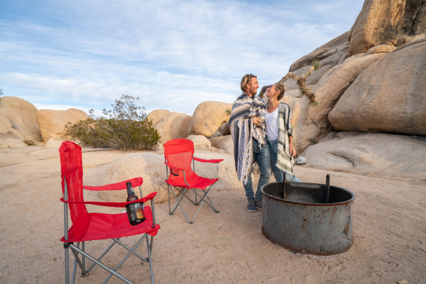 年轻夫妇露营在自然附近的巨石在日落;两个人坐在露营椅上，靠近篝火，享受着夜晚的假期图片下载