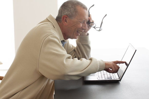 一名男子坐在那里，指着笔记本电脑的屏幕大声笑着。他在屏幕上读到或看到一些非常有趣的东西。我左手拿着一副眼镜。——图片图片下载