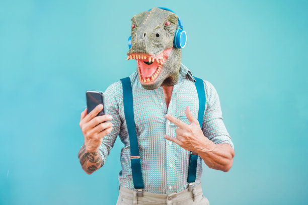 疯狂的纹身男子戴着t-rex面具一边听着音乐一边用智能手机-疯狂的资深男性玩手机应用-科技趋势和时尚概念-专注于脸图片下载
