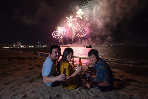 一群朋友手里拿着啤酒瓶，面带微笑，背景是晚上的海滩上五颜六色的烟花图片下载