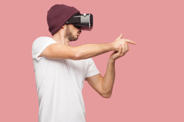 自信的大胡子年轻潮男穿着白衬衫和休闲帽站着，戴着vr玩视频游戏，展示枪唱着手指像射击。图片下载
