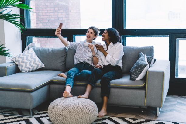 有趣的白人年轻女子与积极的非洲裔美国嬉皮士女孩休息在家里，并在现代智能手机设备上为互联网博客在友好的会议期间自拍照片图片下载