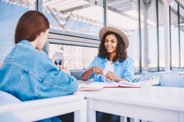 快乐开朗的嬉皮士女孩摆着各种各样的姿势，看着智能手机的摄像头，而女性朋友坐在前面，拍她在准备大学考试的时候一起在街头自助餐厅图片下载