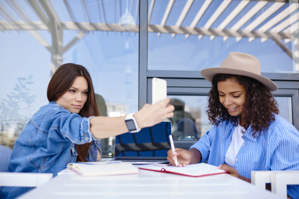漂亮的白人女性在智能手机上自拍，而她的非洲裔美国朋友写作业在笔记本上，时髦的女孩享受空闲时间在咖啡馆露台拍照和做笔记图片下载