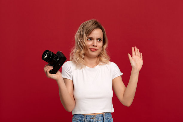 一个金色卷发的女孩的肖像穿着白色t恤站在红色的背景上。模型持有复古相机与姿态下降。图片下载
