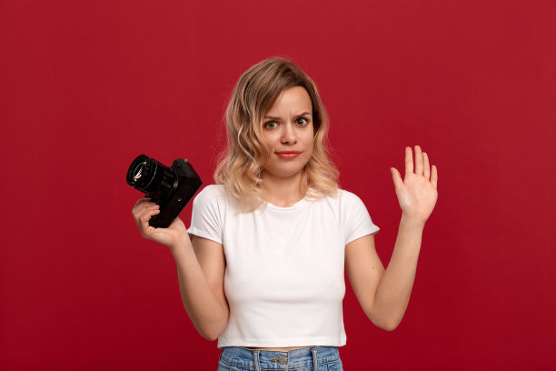 一个金色卷发的女孩的肖像穿着白色t恤站在红色的背景上。模型持有复古相机与姿态下降。图片下载
