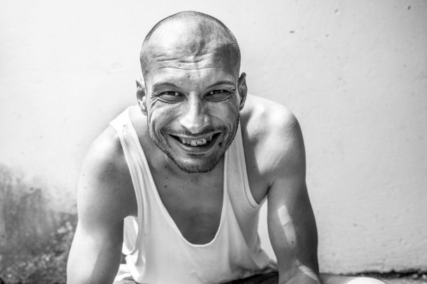 年轻的积极瘦骨嶙峋的厌食症秃头的快乐微笑的流浪汉坐在城市的街道上，在城市或城镇的墙边用大大的微笑看着镜头，流浪汉社会的概念纪录片图片下载