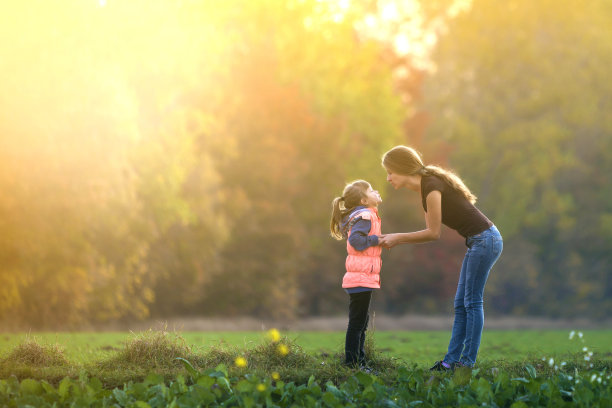 侧面丰满的肖像，年轻苗条迷人的母亲与孩子女孩站在绿色的草地上，在森林树木模糊的背景牵手。图片下载