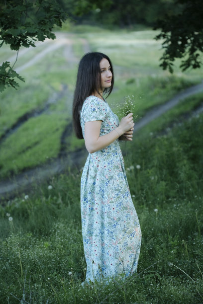 年轻女子手持一束鲜花，站在橡树树冠下郁郁葱葱的林间空地上图片下载