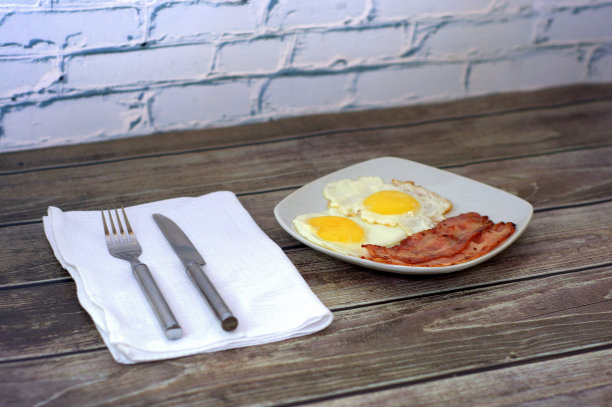 早餐，木桌上放着一个装有煎蛋和熏肉的盘子，旁边放着餐巾和刀叉。特写镜头。图片下载