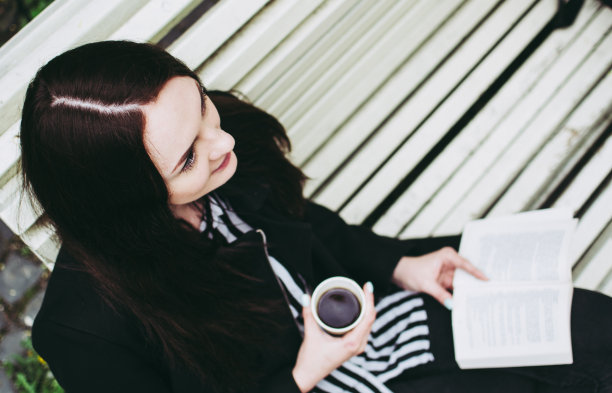 美丽的苍白的脸年轻的黑发阅读一本畅销的书和喝咖啡在一个公园在春天。图片下载