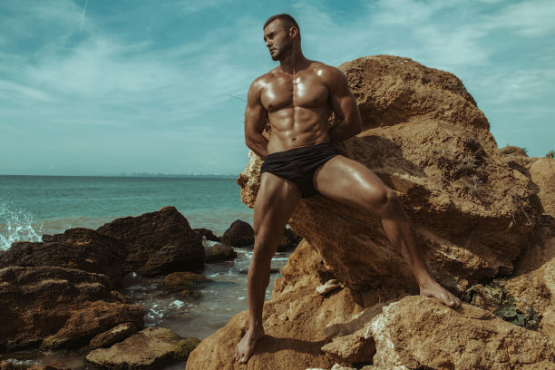 性感的运动肌肉美丽的条纹男模特与白色的毛巾和泳衣在自然的背景与大海和岩石，沙子和天空在海滩上的日落图片下载