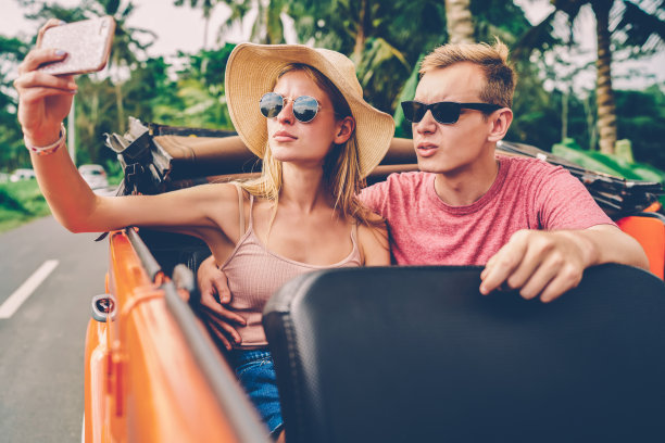 时髦的女孩戴着墨镜用智能手机拍照，和男友自拍分享到网络上，相爱的情侣在公路上租了一辆车，在手机上摆姿势拍摄共同的照片图片下载