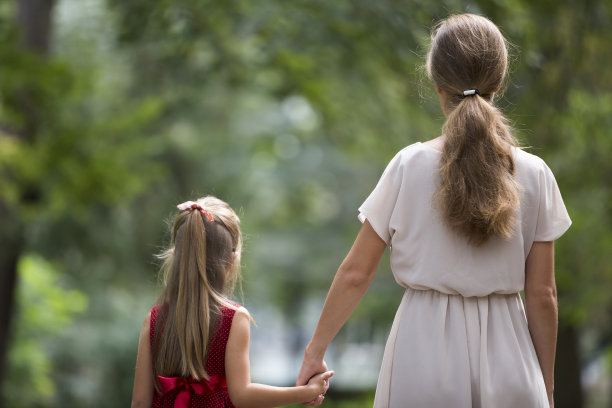 小金发长头发的孩子女孩与苗条苗条的母亲在时尚的服装一起走在温暖的阳光明媚的日子在户外模糊的彩色阳光散景背景手拉手。图片下载