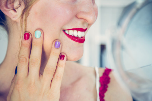 迷人的红唇:金发女郎涂着彩色指甲油看着镜子里的自己。从脸上切下来。图片下载
