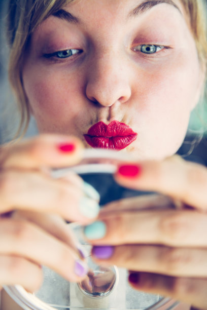 迷人的红唇:金发女郎涂着彩色指甲油看着镜子里的自己。吻的嘴。图片下载