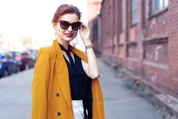 肖像时尚女人戴着太阳镜走在街上。她穿着黄色的夹克，微笑着。图片下载