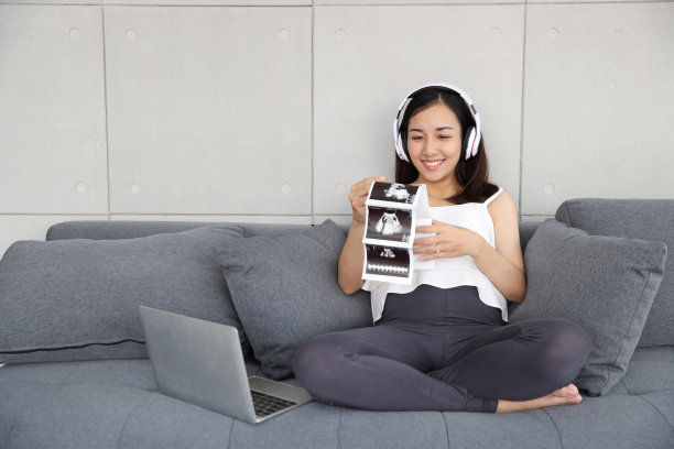 美丽的亚洲孕妇在客厅沙发上用电脑听音乐，同时拿着超声波照片图片下载