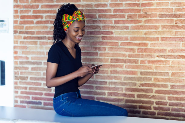 一个快乐的年轻女子坐在墙前的桌子上用手机聊天。图片下载