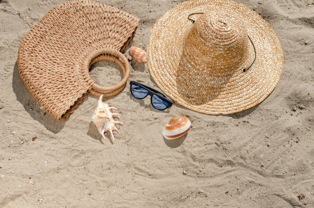 戴着草帽的太阳镜、包和贝壳躺在沙滩上。暑假的场景图片下载