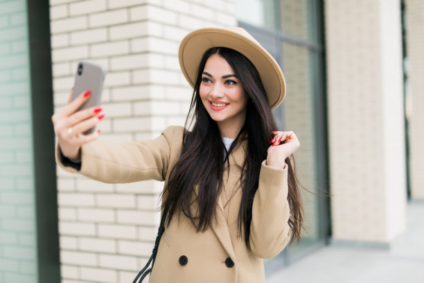 一个年轻漂亮的女人在电话旁自拍。微笑着，走在城市的大街上。去购物。穿着时髦的白色外套。图片下载