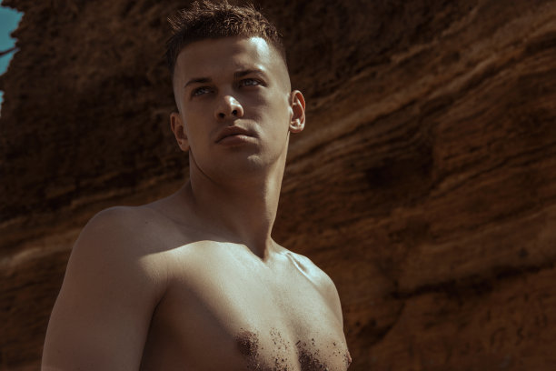 性感的运动肌肉美丽的条纹男模特与白色的毛巾和泳衣在自然的背景与大海和岩石，沙子和天空在海滩上的日落图片下载