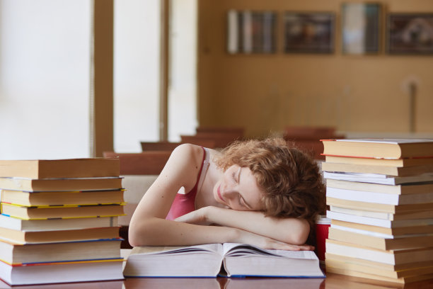 狐狸头发的女学生睡着了，而阅读寻找信息，准备考试，女人躺在桌子上，做作业在大学图书馆。教育、学校的概念。图片下载