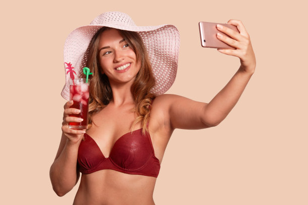 近距离的肖像，有吸引力的白人女人自拍，而持有新鲜的鸡尾酒在手里。美丽的年轻女子在红色泳衣和草帽摆姿势的米色工作室背景。图片下载