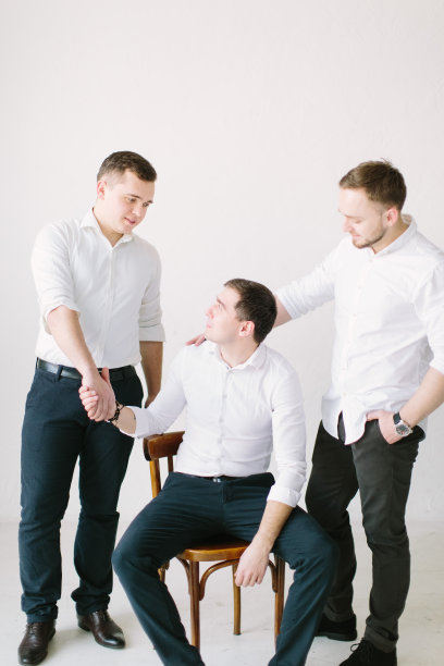 帅气的新郎坐在椅子上摆姿势，伴郎站在他旁边，帮助他早上准备。婚礼准备。黑白照片图片下载