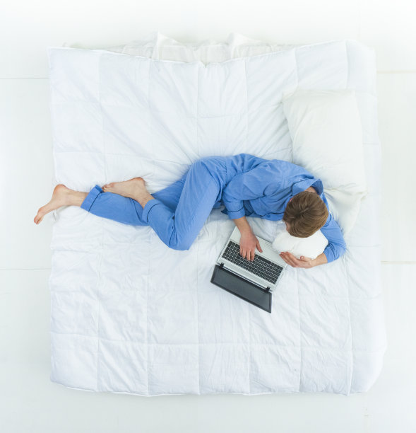 英俊的男人穿着睡衣躺在床上用着他的工作笔记本电脑图片下载