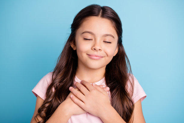 可爱可爱可爱的孩子的近距离照片闭上眼睛触摸的地方手掌的手的胸部感觉休息的灵感愉快她穿着浅色时尚的衣服孤立在蓝色的背景图片下载