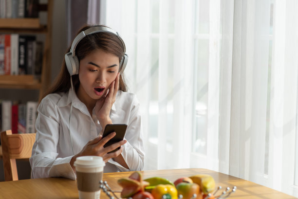 年轻的亚洲女子戴着耳机听着音乐，看着她的智能手机看到坏消息或照片，她震惊困惑的脸上表情图片下载