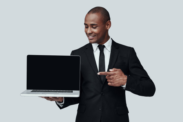 看这里!年轻的非洲男子穿着正装指着笔记本电脑，微笑着站在灰色的背景图片下载