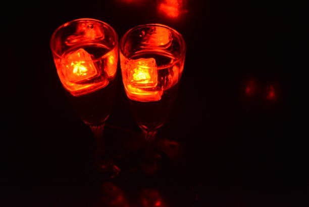 两杯不同寻常的香槟，一杯饮料和鲜红的冰片。在黑色的背景、令人印象深刻的照片和情人节的盛宴上，用塑料冰块照亮每个杯子里的饮料。图片下载