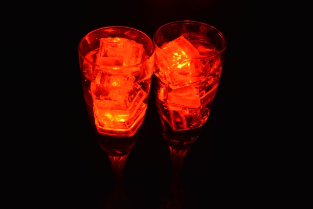 两杯不同寻常的香槟，一杯饮料和鲜红的冰片。在黑色的背景、令人印象深刻的照片和情人节的盛宴上，用塑料冰块照亮每个杯子里的饮料。图片下载