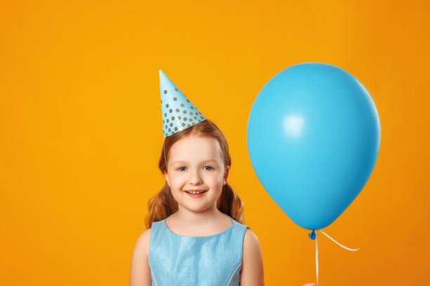 快乐的小女孩庆祝生日。一个孩子拿着一个蓝色的气球，看着镜头。特写肖像上的黄色背景。图片下载