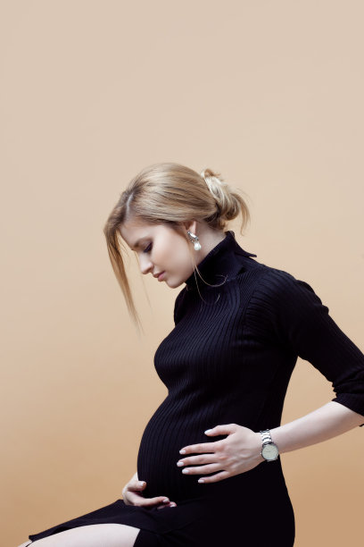 年轻漂亮的孕妇身穿黑色衣服，在米色背景上拥抱她的肚子图片下载