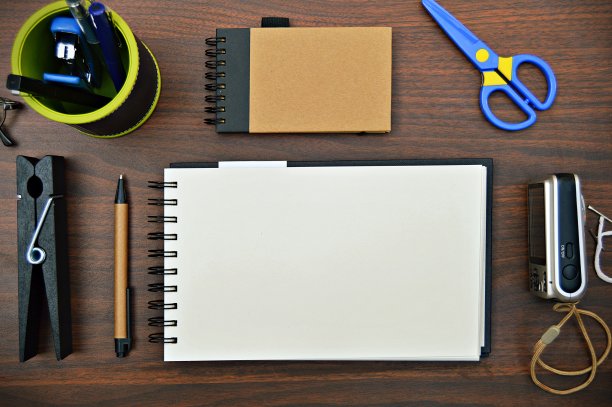 一幅文具的横向照片，如钢笔、日记本、剪刀、笔架、订书机、照相机，放置在一个木制的深棕色水平背景上，非常美观。图片下载