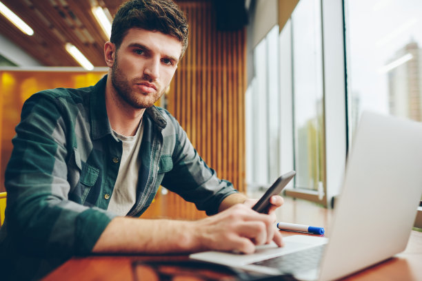 这是一幅男性平面设计师坐在笔记本电脑前，在智能手机上安装4G网络应用程序时看着摄像头的照片。自由职业者在上网本上工作，同时在电话上检查邮件图片下载