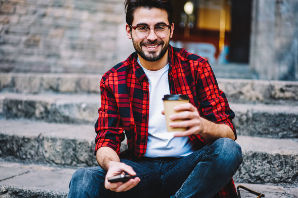 照片中是一名穿着休闲装、戴着眼镜的英俊男子在户外休息，喝着咖啡。照片中是一名兴高采烈的潮男，他坐在街上，手里拿着连接4G网络的智能手机，看着摄像头图片下载