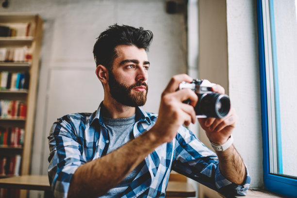 一名留着胡须的职业年轻人，手拿老式相机坐在摄影棚里拍照。创意摄影师聚焦和拍摄现代设备上的照片图片下载