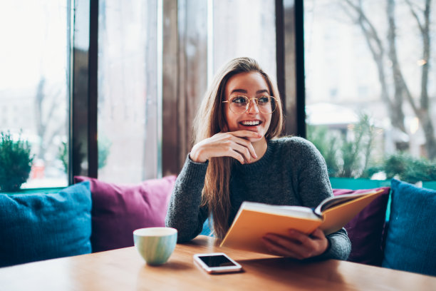 年轻开朗的时髦女孩在时尚的服装拿着笔记本，而看着别处和等待朋友在自助餐厅，微笑的女人在时尚的眼镜享受喜爱的文学室内图片下载