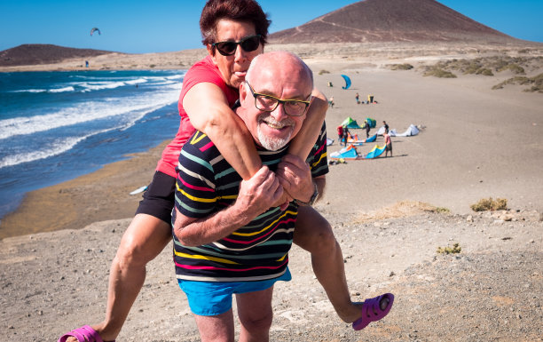 老人肩上扛着妻子。微笑和滑稽的困难。蓝色的天空和海浪为背景的大海。海滩和山图片下载