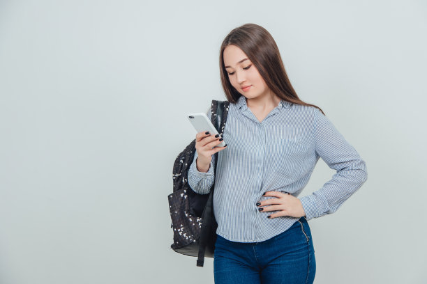 年轻可爱的亚洲学生站着，看着手机，手叉腰，背包挎在肩上。图片下载