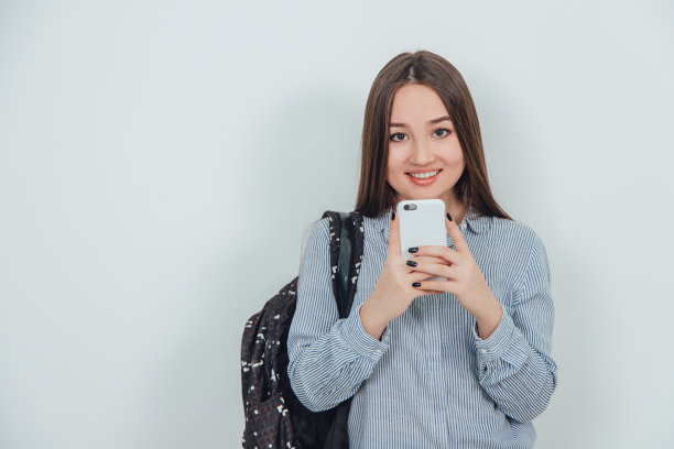可爱的亚洲女学生拿着手机好像在拍照，摆姿势。背包挂在她的肩上。表达的脸。图片下载